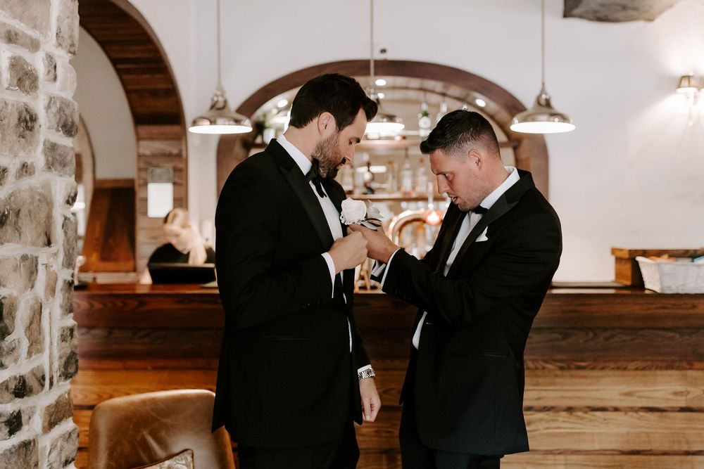 Two people in formal black suits adjusting each other's ties in an elegant indoor venue.