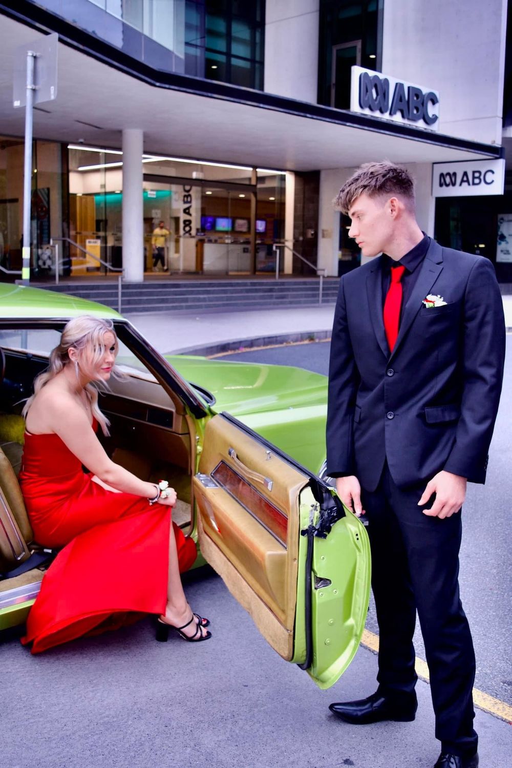 A couple in formal attire poses playfully near a building entrance.