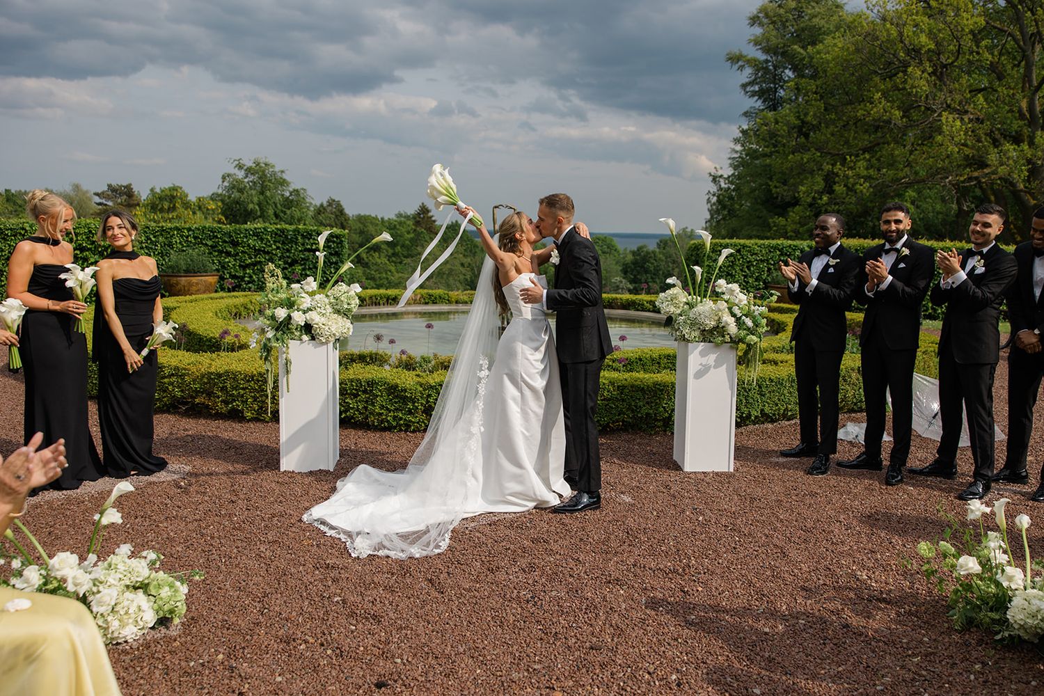Wedding party stands together on a gravel path surrounded by landscaped gardens and cloudy skies.