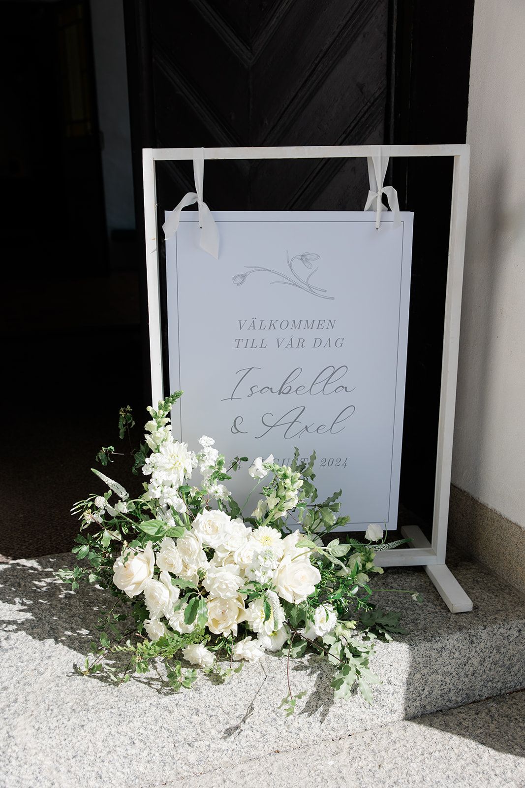 Elegant white floral arrangements with greenery decorate a row of wedding welcome signs on white easels.