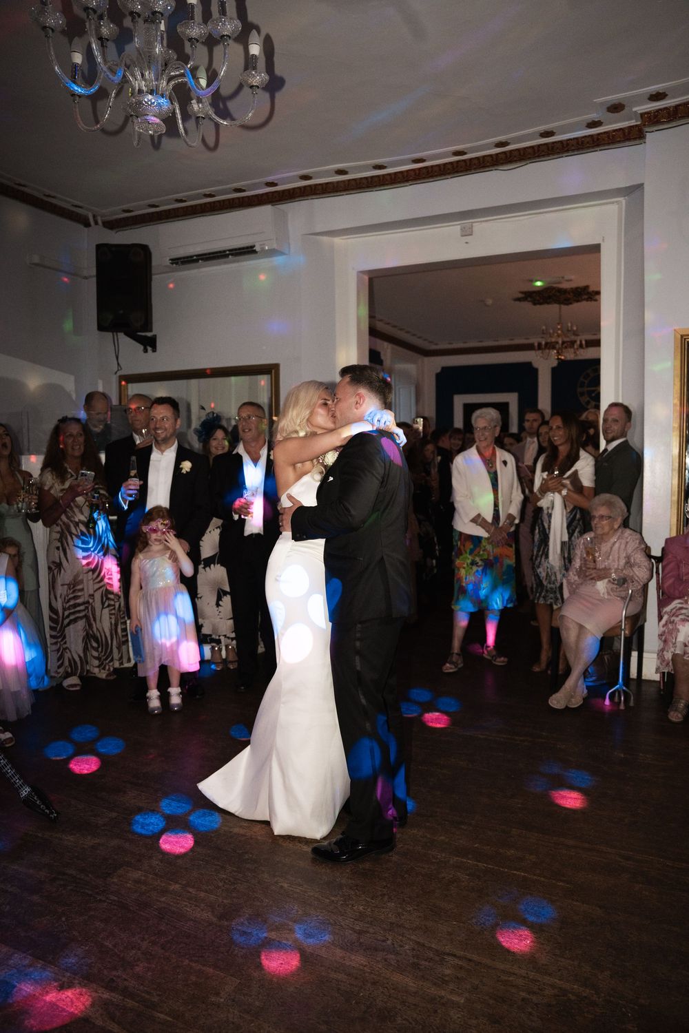 Wedding guests dance on a dark wooden floor under chandeliers with colorful blue and pink lighting effects.