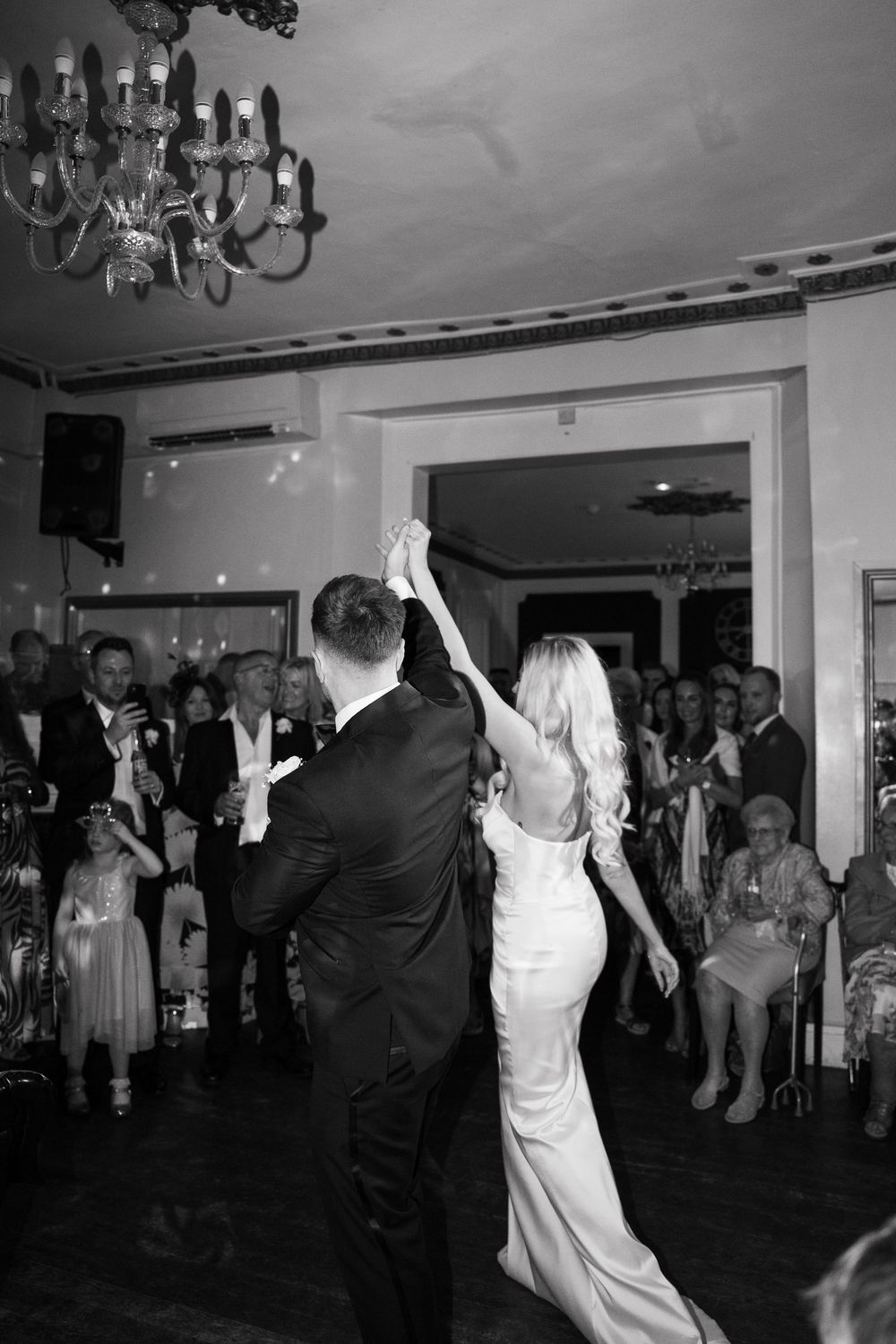A wedding couple dances together on a dark dance floor surrounded by guests at their reception celebration.