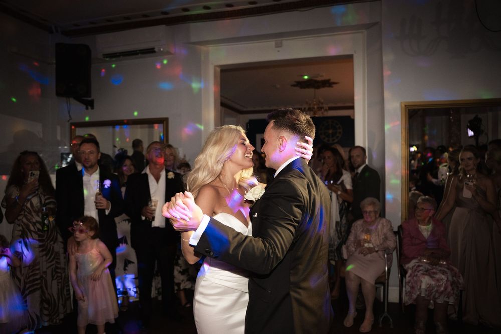 Guests dance and celebrate at a wedding reception with colorful lights and decorations in the background.