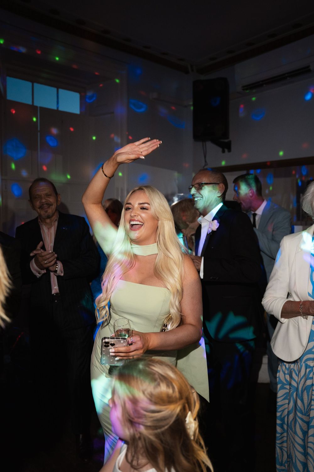 A person in a white dress dances at a party under colorful disco lights with other guests in the background.