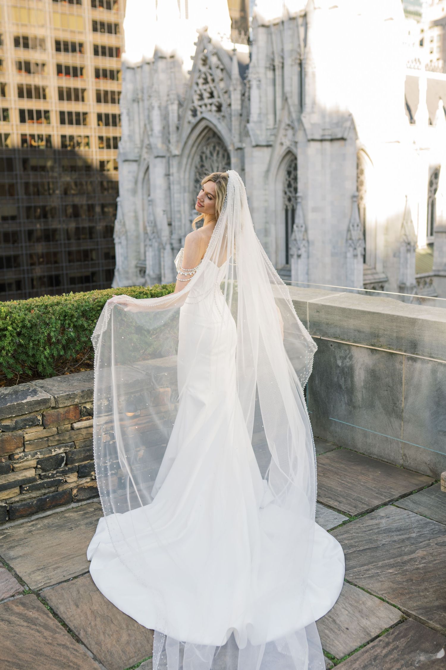 A bride in a flowing white wedding gown and cathedral veil poses on a church rooftop with gothic architecture in the background.