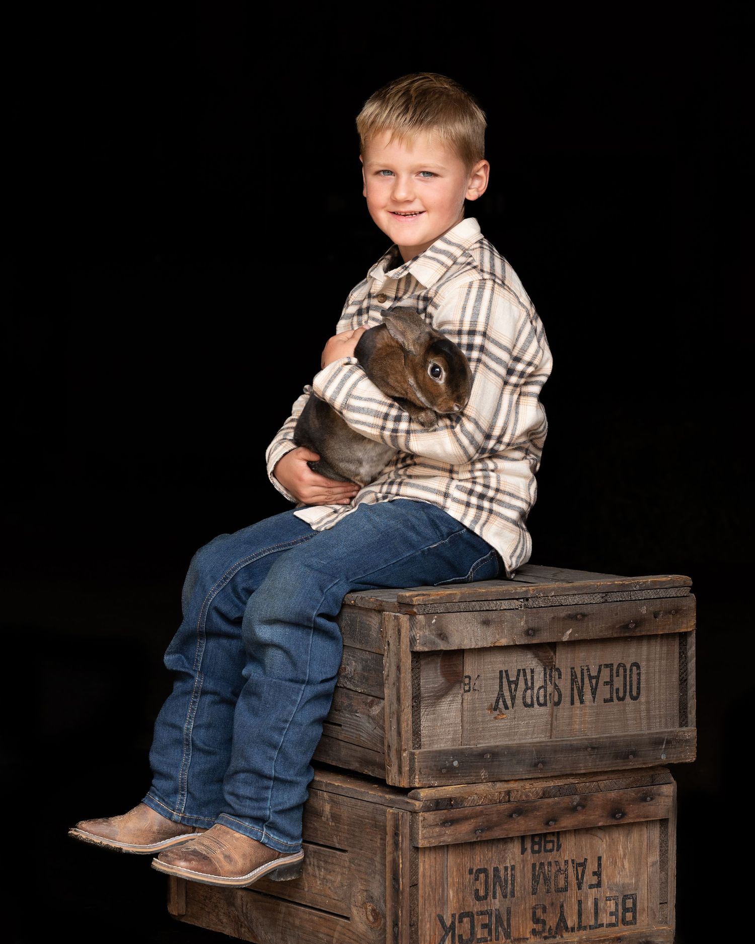 A young child in a plaid shirt and jeans sits atop rustic wooden crates against a black background.