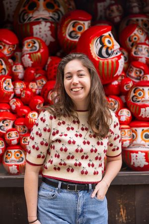 A person in a cherry-print sweater poses in front of traditional red Japanese daruma dolls at a shrine.
