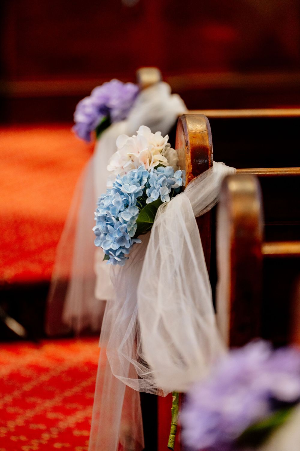 Church pews adorned with white tulle and blue hydrangea floral arrangements for a wedding ceremony.