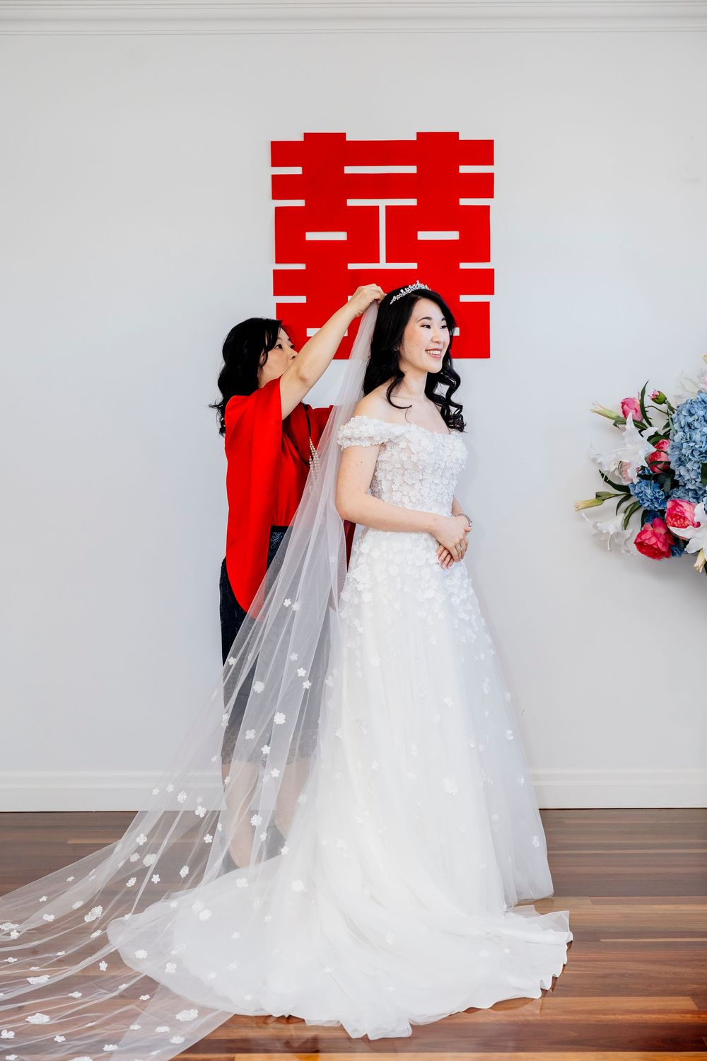 Wedding dress with lace bodice, tulle skirt and delicate floral veil in traditional white against red and gray backdrop.
