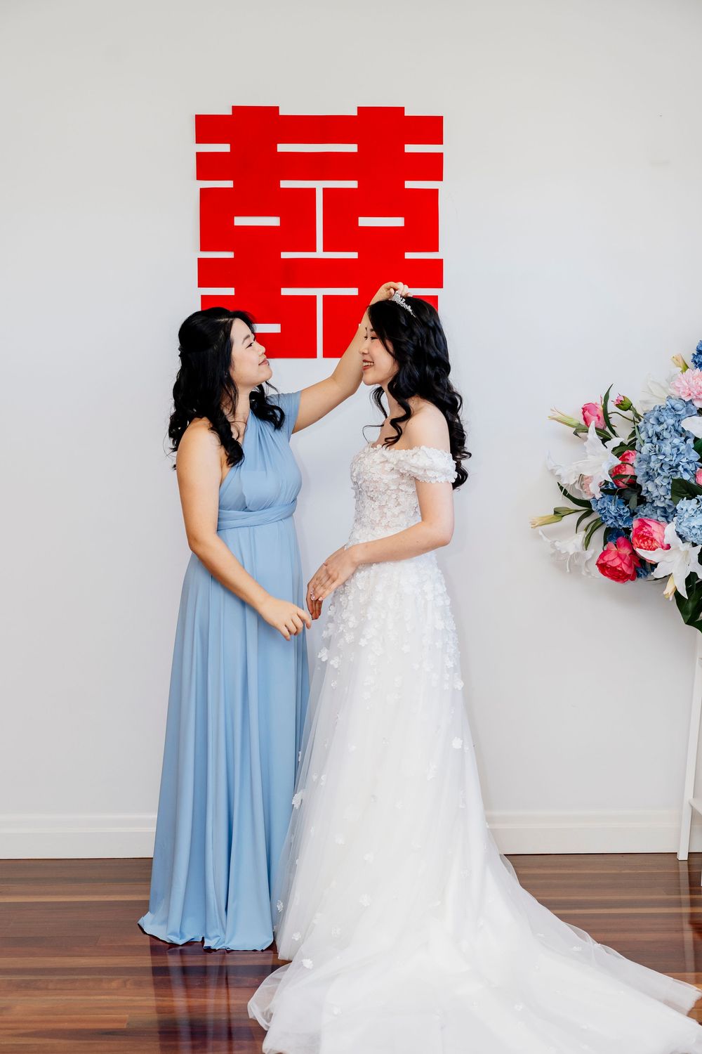 Two people share a joyful moment, one wearing a light blue dress and another in a white wedding gown against a neutral wall.