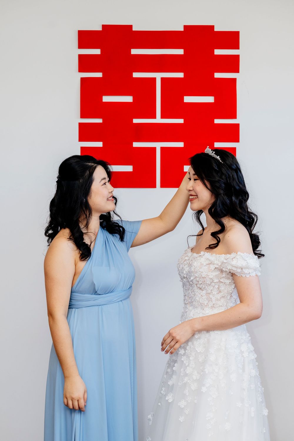 Two people in blue and white dresses share laughter during an intimate photo session against a white and red backdrop.