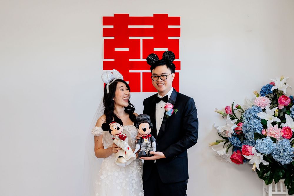 A couple in wedding attire poses together with blue and pink floral arrangements in the background.