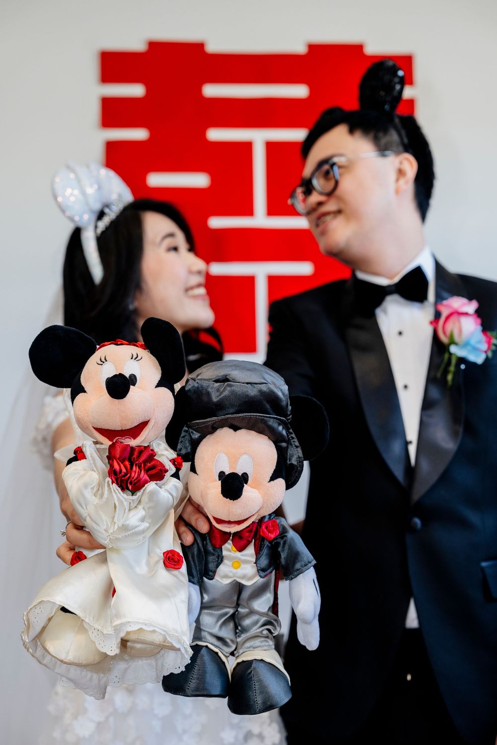 Mickey and Minnie Mouse plush dolls in wedding attire displayed in front of Chinese double happiness symbol.
