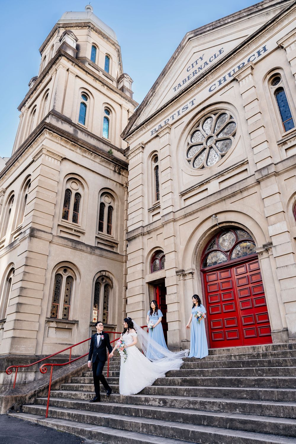 Wedding couple descends grand stone steps of historic church building with dramatic red doors.