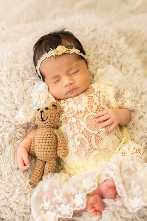 Sleeping newborn baby girl in yellow lace dress with floral headband cuddling a crocheted teddy bear on fluffy white blanket.