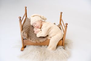 A teddy bear rests on a white fluffy blanket near a wooden four-poster bed in a neutral-toned photography setup.