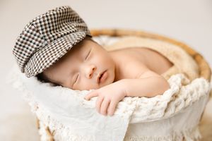 Sleeping newborn wrapped in cream blanket wearing checkered newsboy cap resting in round woven basket.