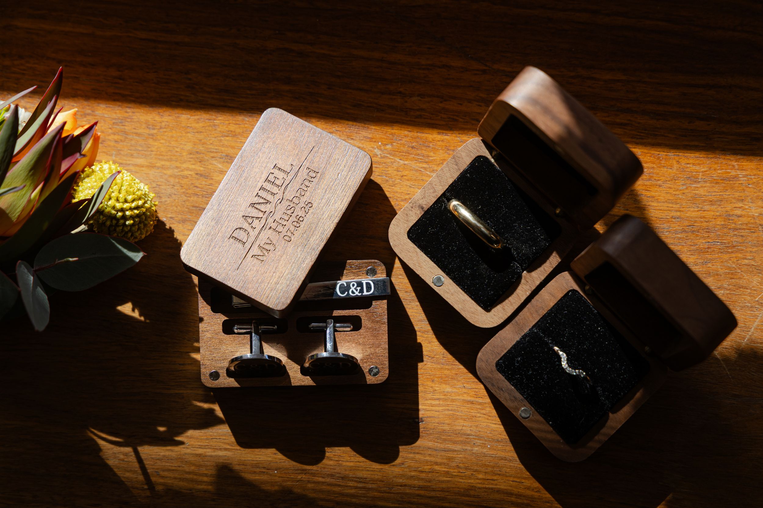 Grooms Prep detail shots of rings, boutonniere, cuff links across a dark wooden surface in dramatic lighting.
