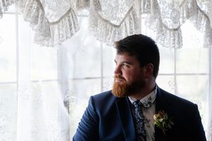A bearded person in a navy suit with boutonniere gazes thoughtfully by lace curtained window.