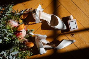 Wedding details with elegant white bridal shoes and pink tulip bouquet arranged on a wooden floor in natural light.