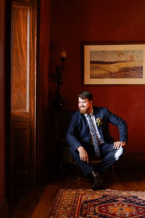 moody interior shots of groom in a navy suit sitting in an arm chair with ornate rugs on a hardwood floor with red walls.