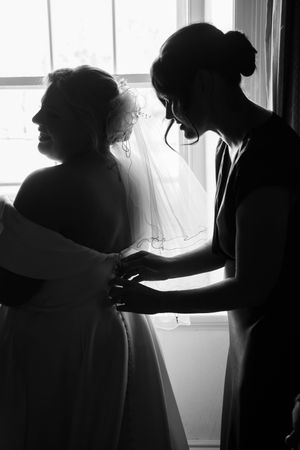 Black and white silhouettes of people in wedding attire preparing by a bright window.