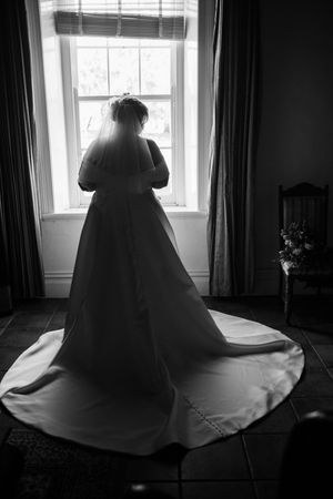 Dramatic black and white photo of bride by window showing off her wedding dress silhouette