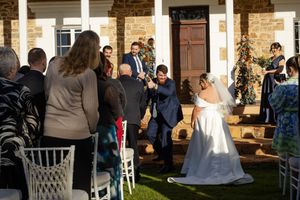 A wedding ceremony takes place on a lawn in front of a stone building with guests seated in white chairs.