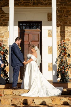 bride and groom standing hand in hand under a rustic stone porch entrance decorated with floral arrangements for a wedding ceremony.