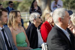 Wedding guests dressed in formal attire gather outdoors during a ceremony on a sunny afternoon.