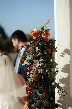 Orange and white floral arrangements frame a sunlit wedding ceremony scene with white columns.