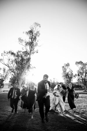 Black and white silhouette of wedding party running across field at sunset.