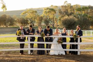 Wedding group poses lined up along white fence during beautiful sunset in countryside.