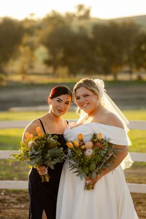Wedding attendants hold autumn-themed bouquets while sharing a joyful moment in warm evening light.
