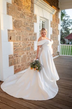 Beautiful white wedding dress shown in detail against rustic stone wall backdrop on wooden porch.