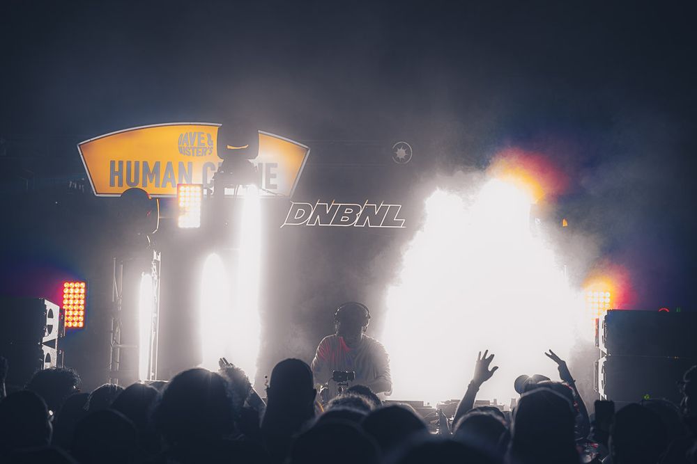 Crowd of concert-goers waves hands in the air as bright stage lights illuminate a dark performance venue.