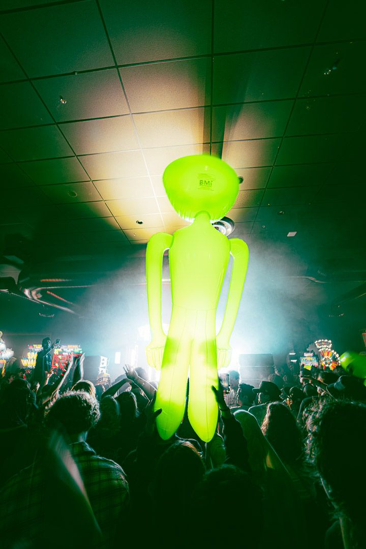 Large neon green inflatable figure glows above crowd at indoor concert venue.