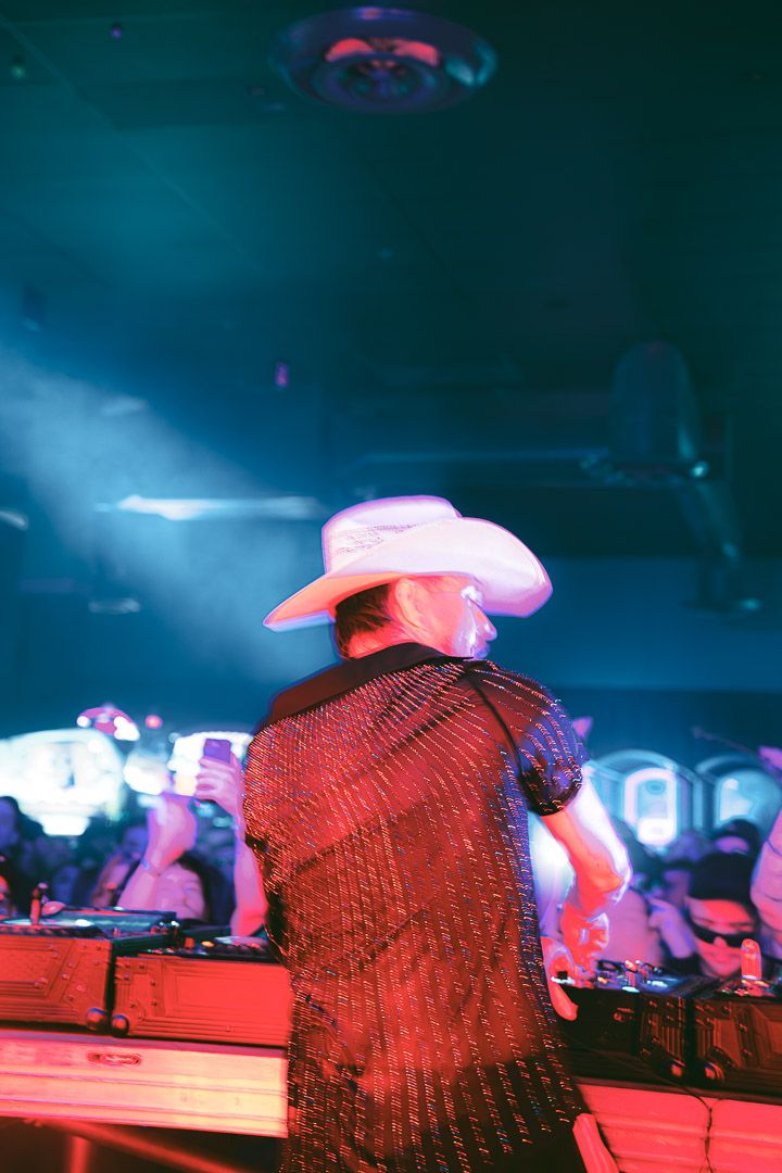 DJ wearing a cowboy hat performs at a nightclub with colorful lighting effects in the background.