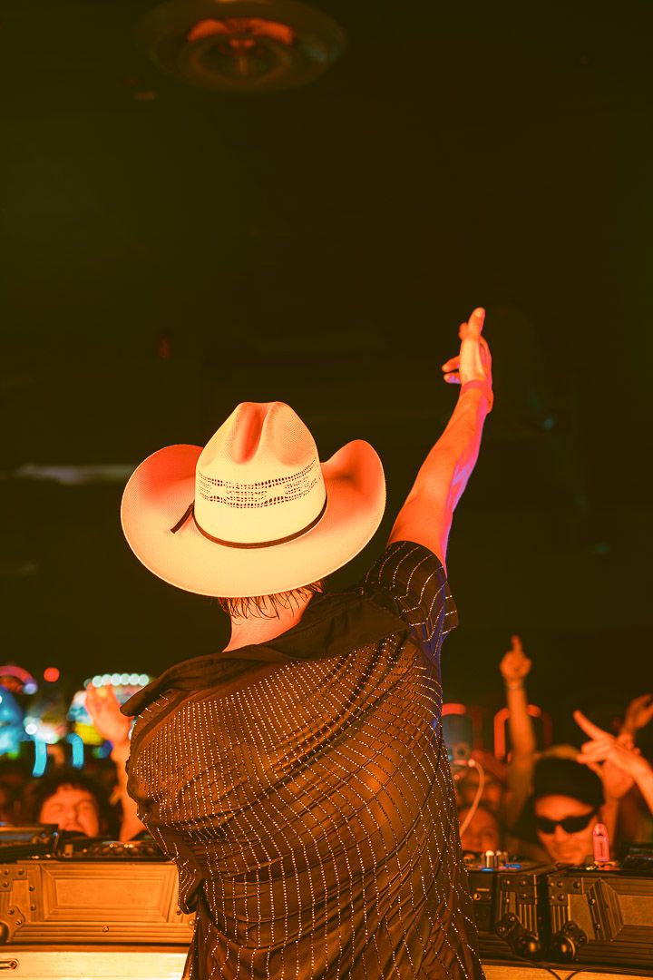 Country music concert crowd silhouette with cowboy hats glowing orange under stage lights against dark backdrop.