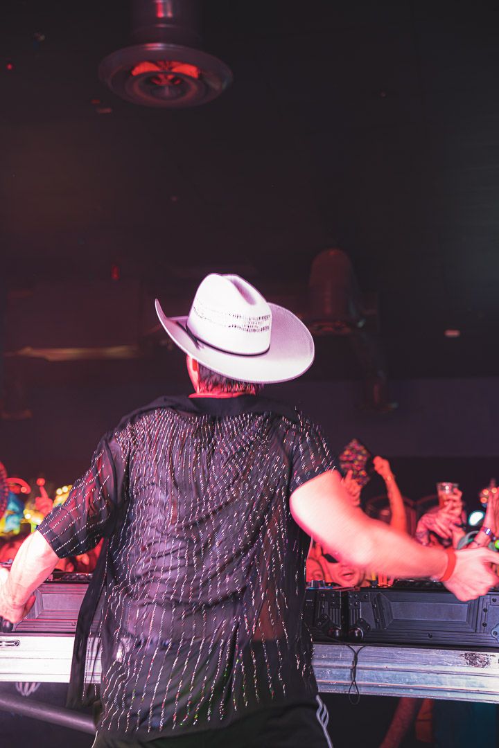DJ in cowboy hat and dark shirt performs at nightclub with red lighting and crowd in background.