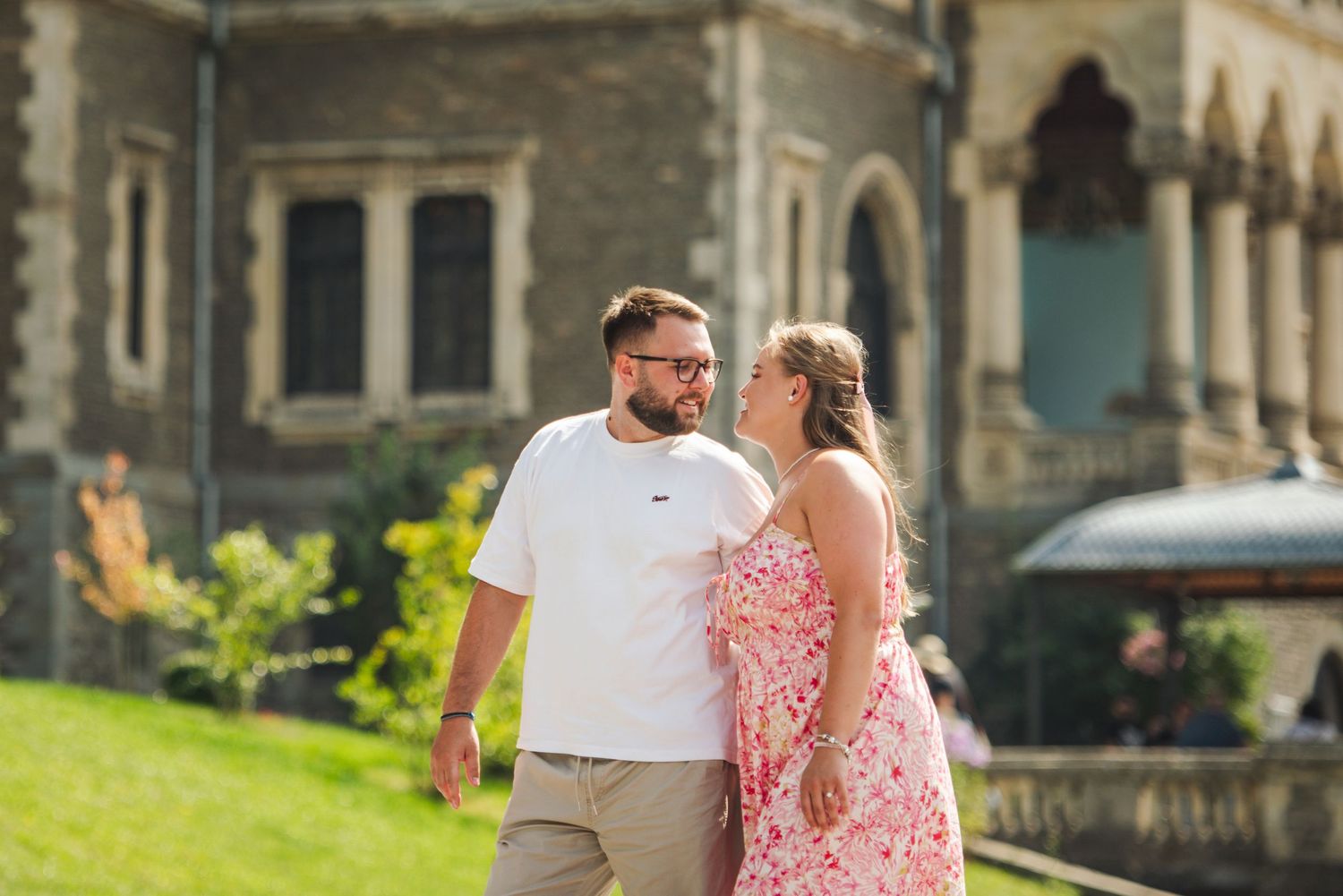 Couple in pink dress and white shirt walking together on a grassy path near stone buildings.