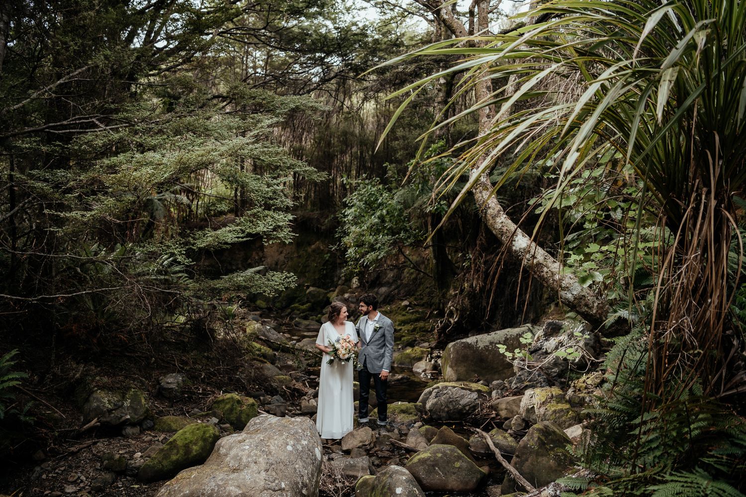 Newlyweds stand on rocks in a river in a chasm amongst NZ native foliage during their Wairarapa heli-elopement by August and Vine