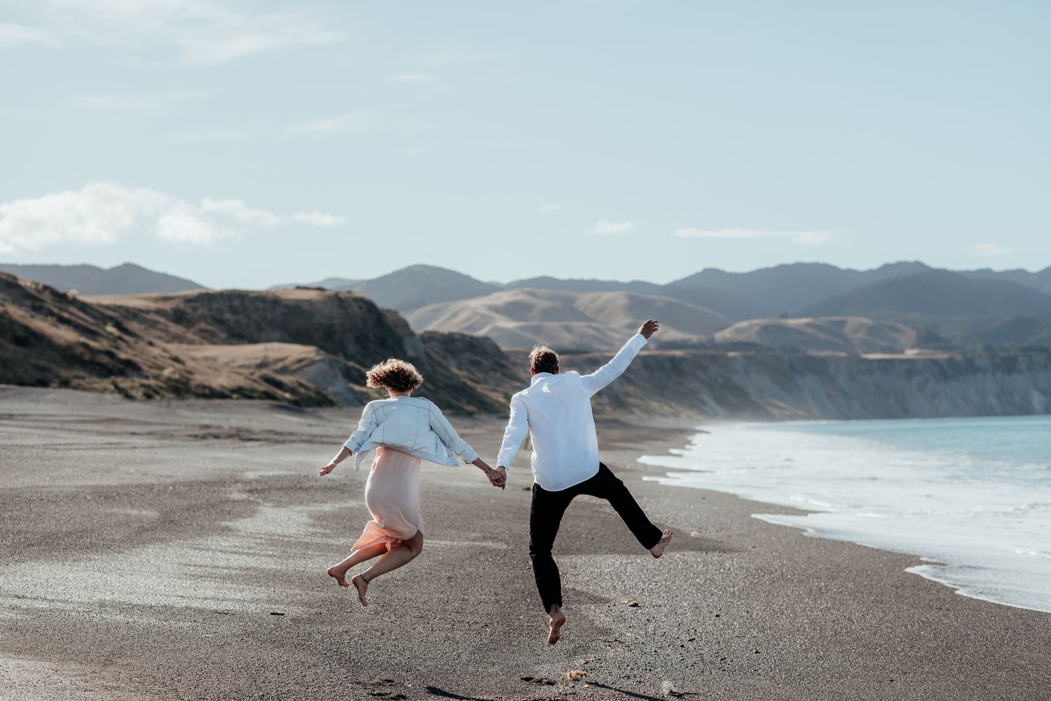 A just married couple jump joyfully on scenic Whangaimoana beach Wairarapa with mountains and blue ocean in the background by August & Vine