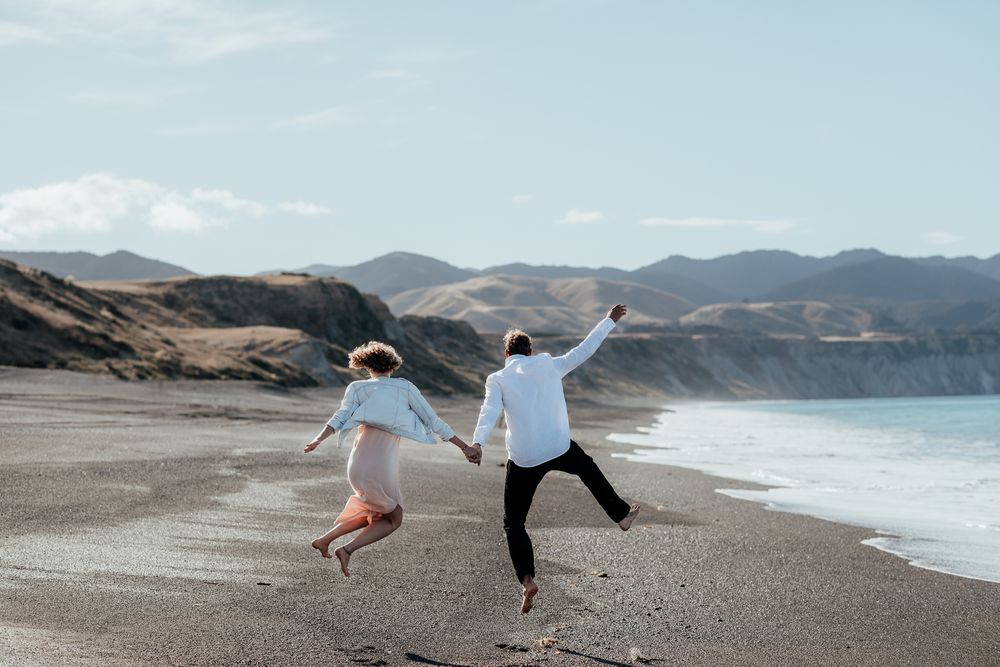 A just married couple jump joyfully on scenic Whangaimoana beach Wairarapa with mountains and blue ocean in the background by August & Vine