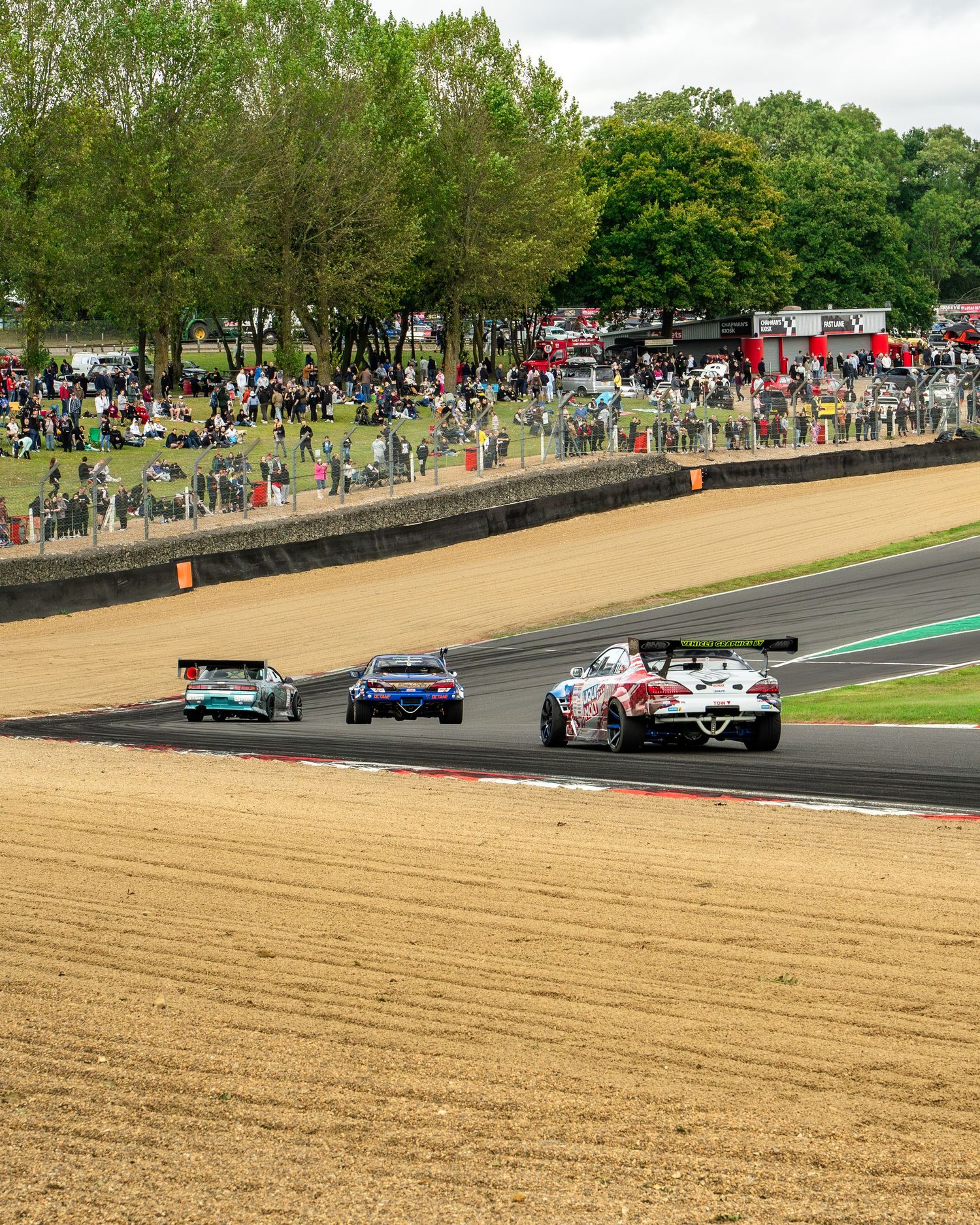 Spectators line the edge of a race track with a groomed sand trap visible in the foreground.