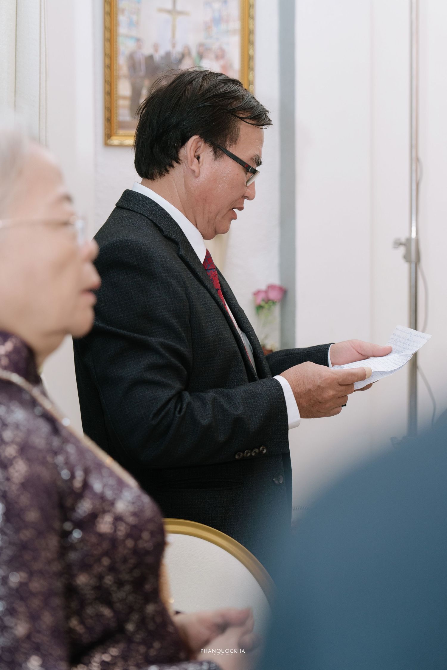 Formal man in black suit reads paper at a traditional Vietnamese wedding ceremony beside elderly woman.