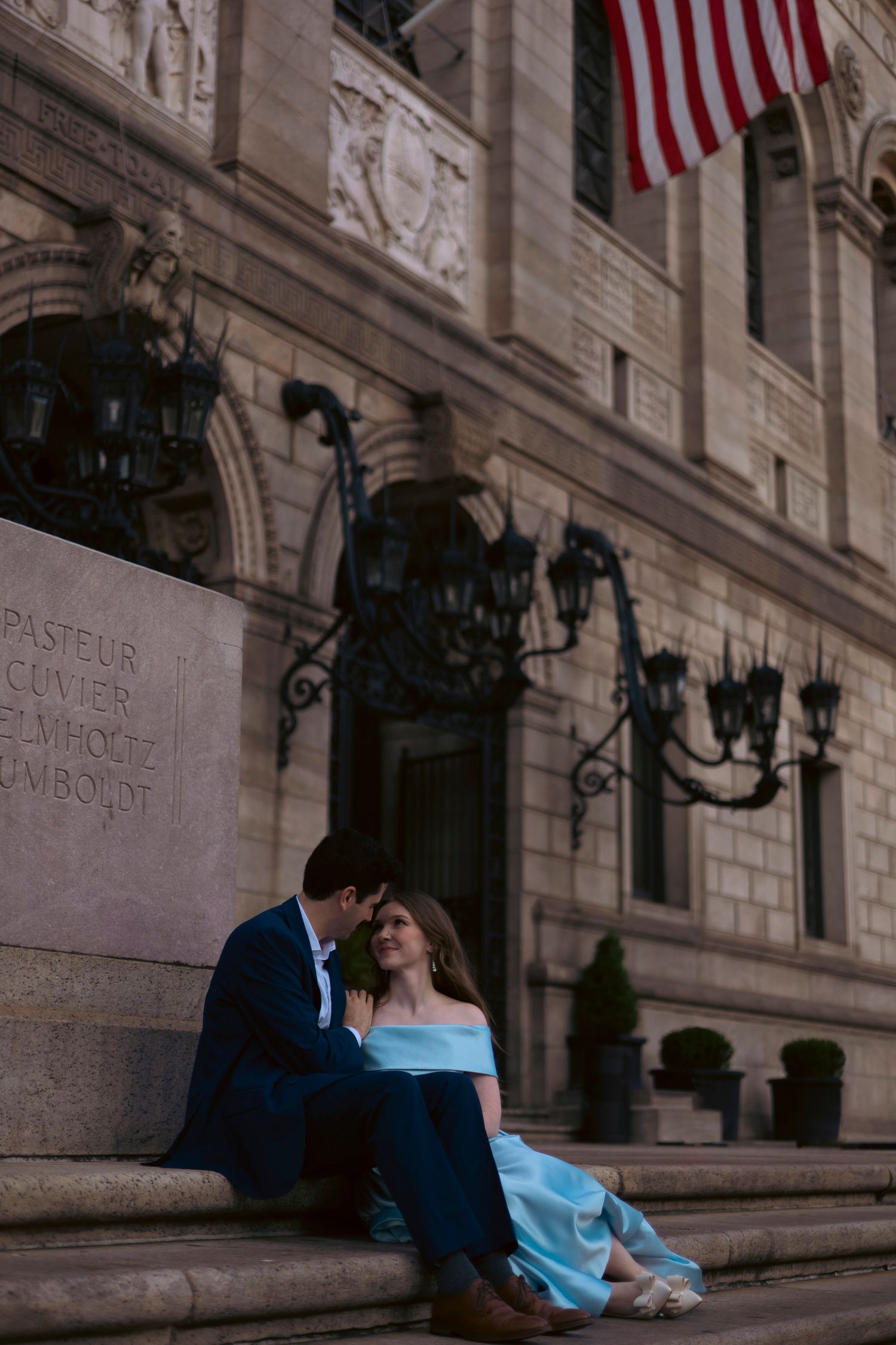 Romantic couple in formal attire sits on stone steps of historic building with American flag above.