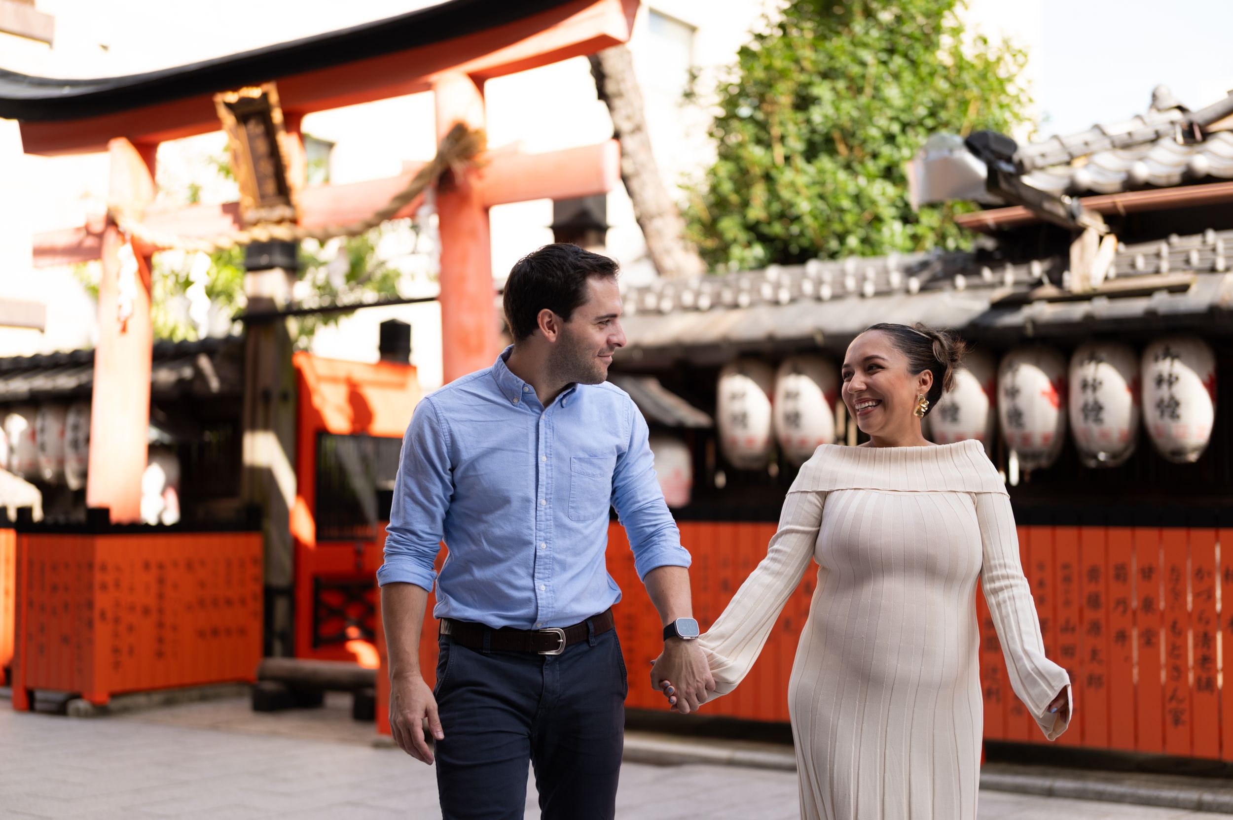 Couple holding hands and smiling at each other in front of a traditional Japanese torii gate with lanterns.