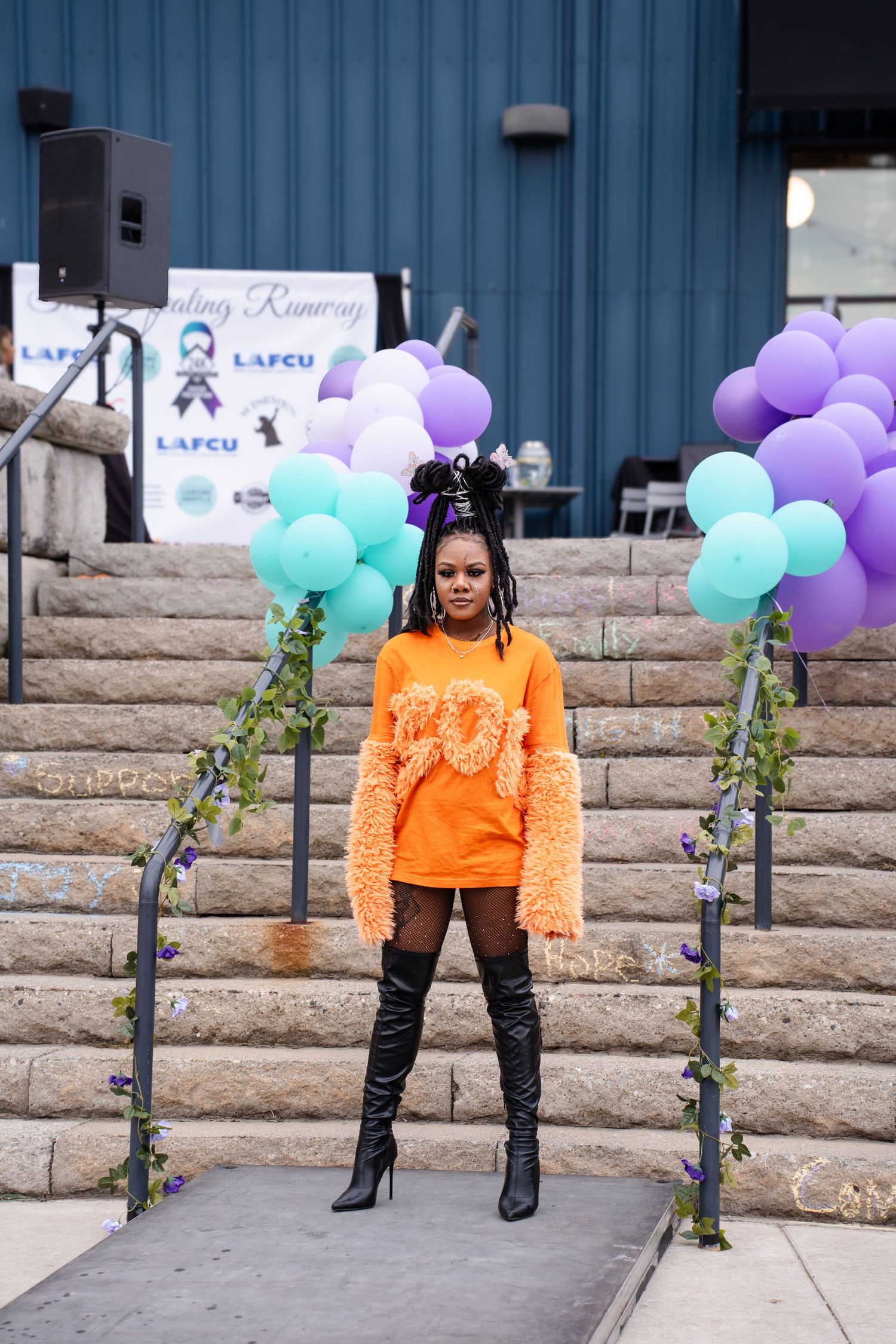 Person in bright orange sweater poses confidently with purple and teal balloons against stone wall backdrop.