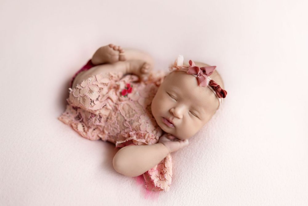 A sleeping baby wearing pink lace outfit lies peacefully on a soft pink background.